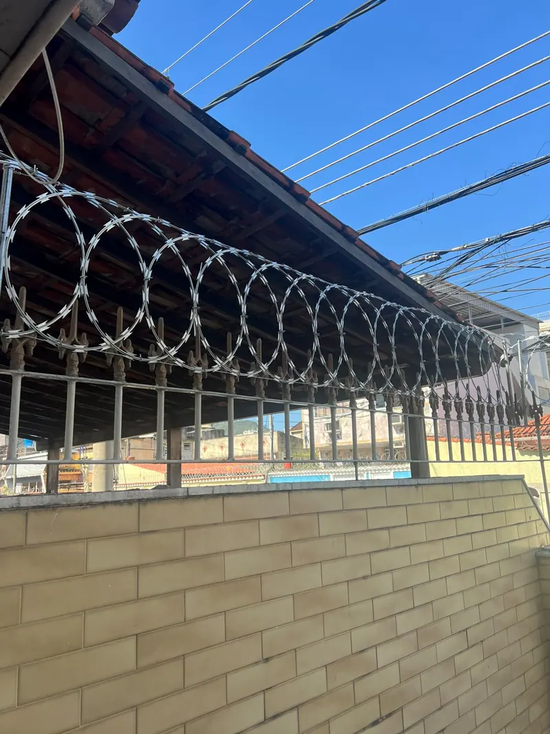 Concertina instalada no alto de um gradil de ferro, embaixo do telhado de uma casa, com o céu azul ao fundo