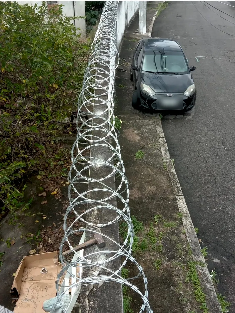 Concertina sendo instalada no alto de um muro, vista de cima, com um martelo e uma caixa de papelão ao lado